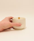 Hand holding a small white ceramic container on a wooden surface with a blurred background