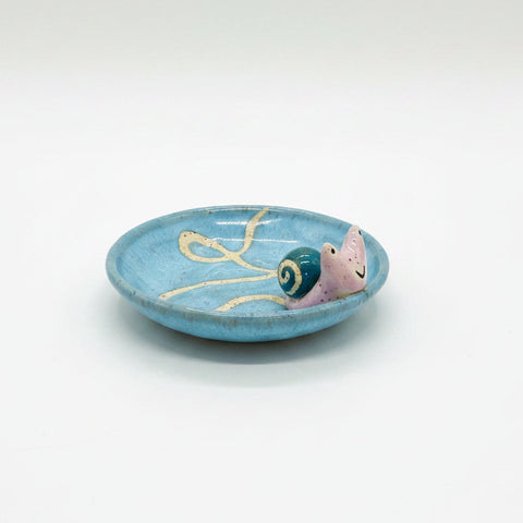 Ceramic snail on a blue plate on a wooden table with a laptop and snacks in the background.