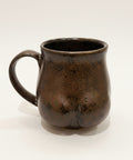 Black ceramic mug shaped like a butt on a wooden surface with an Apple product in the background.