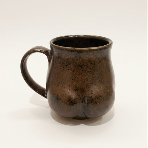 Black ceramic mug shaped like a butt on a wooden surface with an Apple product in the background.
