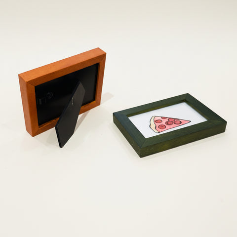 Two small picture frames on a wooden surface, one with a black background and the other with a pizza illustration.