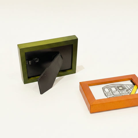 Small green picture frame on a wooden surface with another frame displaying a drawing of a train.