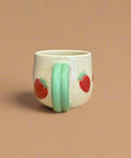 White mug with green handle and strawberry designs on a cardboard surface