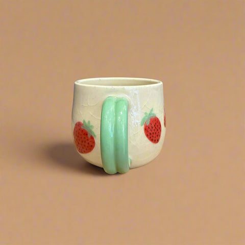 White mug with green handle and strawberry designs on a cardboard surface