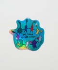 Holographic sticker with cartoon characters and 'be kind to all kinds' text on a wooden surface