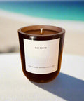 Candle in a dark brown container with a white label on a white surface with ocean in the background