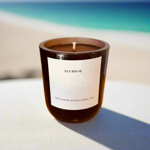 Candle in a dark brown container with a white label on a white surface with ocean in the background