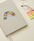 A die-cut holographic sticker with the text 'Love Yourself First' in white letters, surrounded by colorful illustrations of fruit and a rainbow, placed on a light wood surface.
