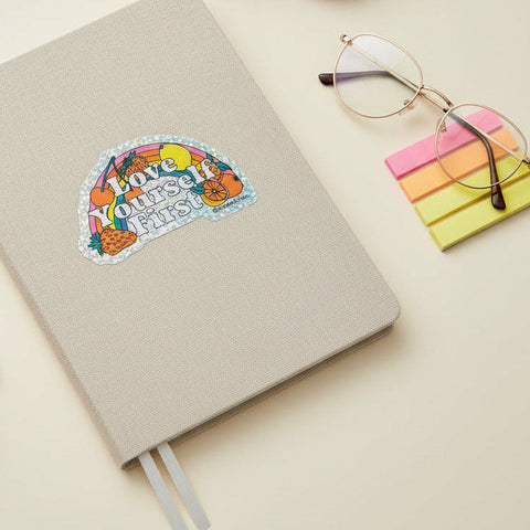 A die-cut holographic sticker with the text 'Love Yourself First' in white letters, surrounded by colorful illustrations of fruit and a rainbow, placed on a light wood surface.
