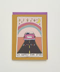 A greeting card with a whimsical illustration of a pink car driving down a road under a rainbow and crescent moon, placed on top of a purple envelope on a light wood surface.