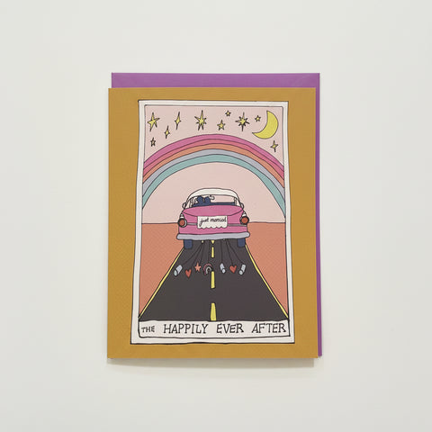 A greeting card with a whimsical illustration of a pink car driving down a road under a rainbow and crescent moon, placed on top of a purple envelope on a light wood surface.