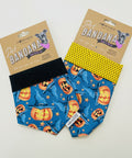 Two handmade pet bandanas displayed on a light wood surface. One bandana is blue with a jack-o'-lantern and star pattern, featuring a black elastic strap.