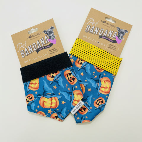 Two handmade pet bandanas displayed on a light wood surface. One bandana is blue with a jack-o'-lantern and star pattern, featuring a black elastic strap.