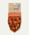 A handmade pet bandana for dogs, featuring an autumn leaf pattern in orange, brown, and black, with a solid rust-orange patterned cuff. The bandana is attached to a brown kraft paper header card with a dog illustration, displayed flat on a light wood background.