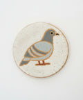 A round ceramic coaster featuring a stylized pigeon illustration in muted blue-gray and brown on a speckled off-white background, placed on a light wood tabletop.