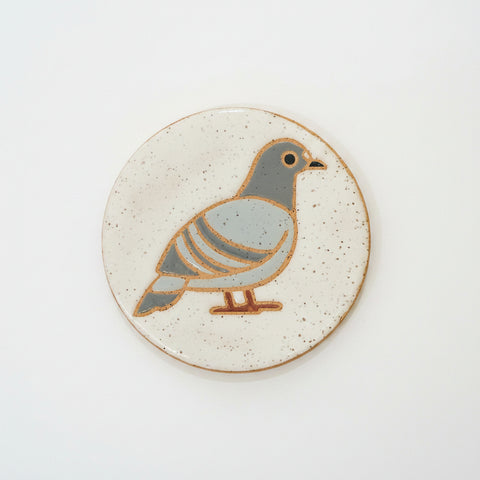 A round ceramic coaster featuring a stylized pigeon illustration in muted blue-gray and brown on a speckled off-white background, placed on a light wood tabletop.