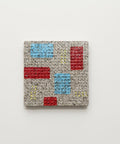 A square coaster with a modern geometric pattern of red and light blue blocks on a beige background, placed on a light wood tabletop.