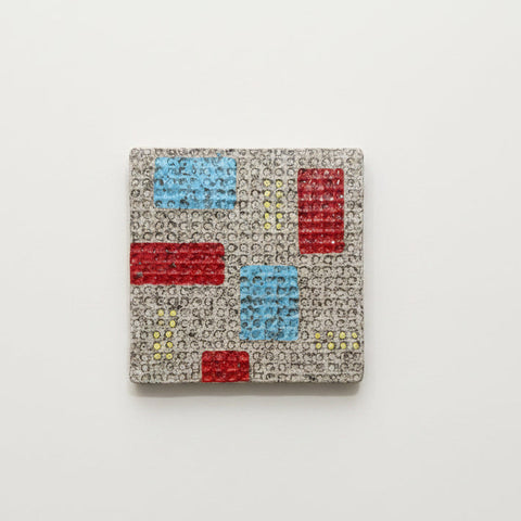 A square coaster with a modern geometric pattern of red and light blue blocks on a beige background, placed on a light wood tabletop.