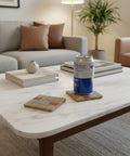 A silver and blue beverage can sits on a woven square coaster on a wooden table, with another patterned coaster nearby and a blurred shop interior in the background.