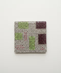 A square coaster with a geometric pattern of green, burgundy, and red diamond shapes on a beige textured background, placed on a wooden table.