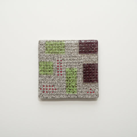 A square coaster with a geometric pattern of green, burgundy, and red diamond shapes on a beige textured background, placed on a wooden table.