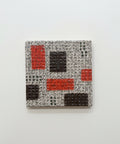 A ceramic coaster featuring an abstract geometric pattern of red, dark brown, and orange blocks on a textured gray background, placed on a light wood surface.