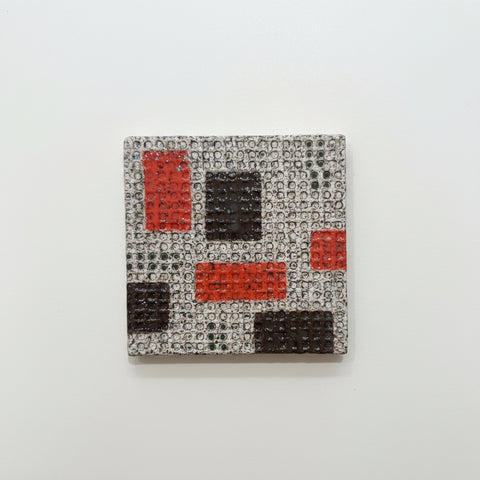 A ceramic coaster featuring an abstract geometric pattern of red, dark brown, and orange blocks on a textured gray background, placed on a light wood surface.