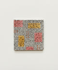 A square coaster with a colorful geometric patterns in pink, yellow, and orange, placed on a wooden tabletop.