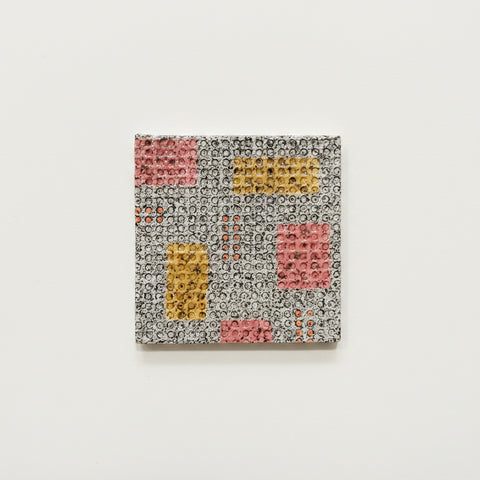 A square coaster with a colorful geometric patterns in pink, yellow, and orange, placed on a wooden tabletop.