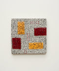A square ceramic coaster with a textured speckled gray base and geometric color blocks of orange, dark red, and light red, shown on a light wood tabletop.
