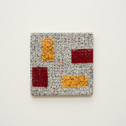 A square ceramic coaster with a textured speckled gray base and geometric color blocks of orange, dark red, and light red, shown on a light wood tabletop.