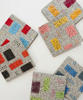 Five square ceramic coasters with colorful geometric blocks, arranged on a light wood tabletop.