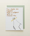 A white greeting card with a hand-drawn stick figure holding a yellow French fry and the text 'You make me happier than a pigeon with a French fry.' placed on top of a sage green envelope.