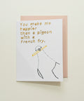 A white greeting card with a humorous quote and a simple drawing, placed on a light pink envelope over a wooden surface.