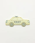A sticker shaped like a vintage taxi car. The sticker is pale yellow-colored with blue stitched outlines and details, and a small sign on top reads 'TAXI'.