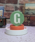 Decorative item with a green circle and white 'G' on a white base, set against a rustic background with a brick wall and vintage suitcase.