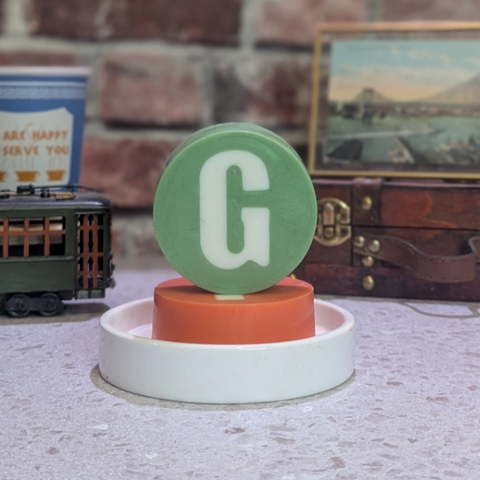 Decorative item with a green circle and white 'G' on a white base, set against a rustic background with a brick wall and vintage suitcase.