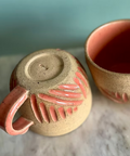 Two ceramic mugs with pink handles and decorative elements on a marble surface.
