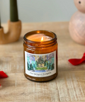 Candle in an amber jar with a label on a wooden surface, surrounded by decorative items.