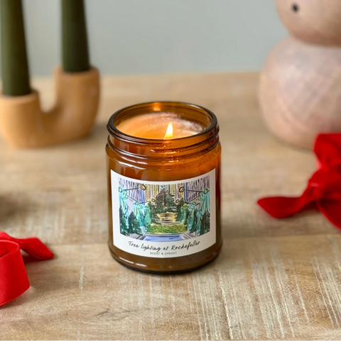 Candle in an amber jar with a label on a wooden surface, surrounded by decorative items.