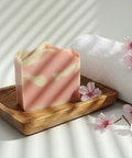 Pink soap bar on a wooden tray with cherry blossoms and a white towel in the background