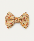 A small fabric bow tie with an autumn leaf pattern in orange, red, and green on a beige background, displayed on a wooden surface against a white wood paneled backdrop.