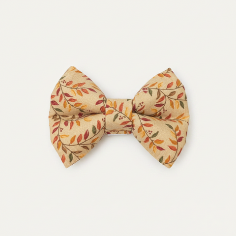 A small fabric bow tie with an autumn leaf pattern in orange, red, and green on a beige background, displayed on a wooden surface against a white wood paneled backdrop.