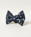 A black fabric bow tie with a celestial print of white and gold crescent moons, stars, and sparkles, displayed on a light wood block against a white wood plank background.