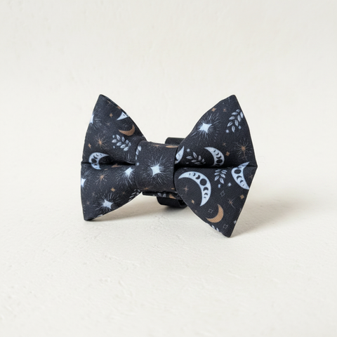 A black fabric bow tie with a celestial print of white and gold crescent moons, stars, and sparkles, displayed on a light wood block against a white wood plank background.
