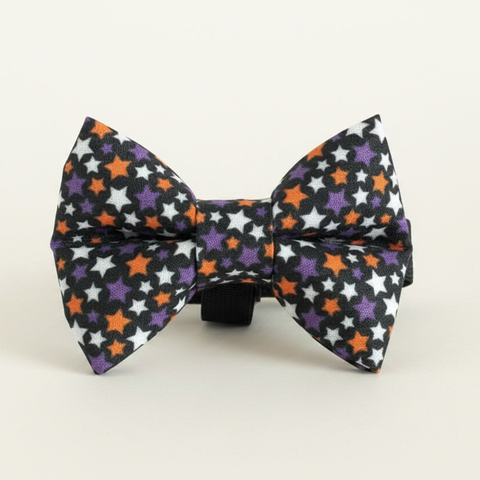 A small bow tie with a Halloween star pattern, featuring black, white, purple, and orange stars on a black background, displayed on a wooden block over a whitewashed wood surface.
