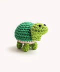A crocheted turtle toy in shades of green, resting on a clear glass cylinder against a plain light background.