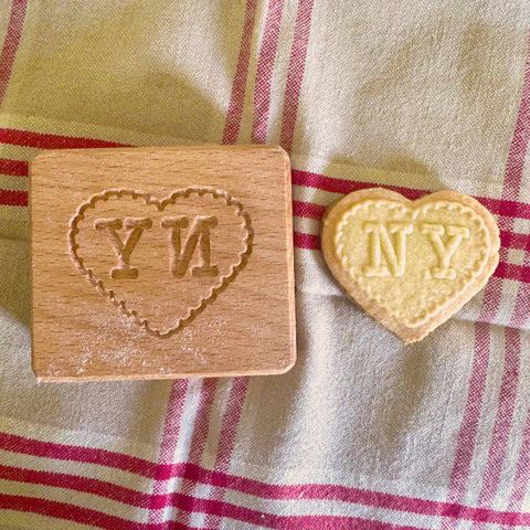 Wooden heart-shaped cookie stamp with 'NY' letters next to a baked heart-shaped cookie with 'NY' letters on a checkered fabric background.