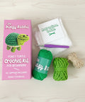 The Pudgy Rabbit Pokey Turtle Crochet Kit shown with its pink packaging box, green yarn, beige yarn, a purple crochet hook, and an instruction booklet on a white background.