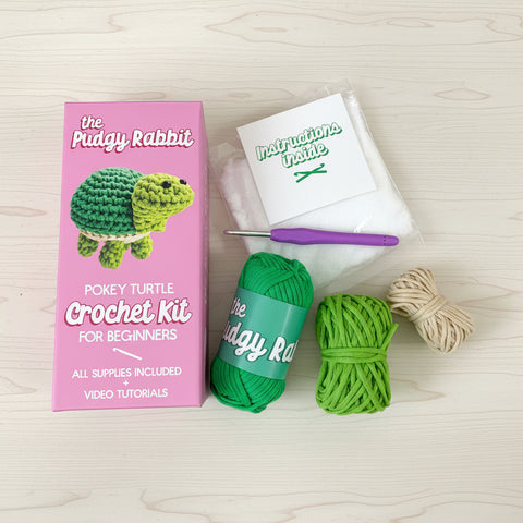 The Pudgy Rabbit Pokey Turtle Crochet Kit shown with its pink packaging box, green yarn, beige yarn, a purple crochet hook, and an instruction booklet on a white background.